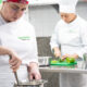 Duas alunas do curso de Gastronomia da Unoeste preparam alimentos durante aula prática em cozinha profissional, enquanto uma mexe uma panela e a outra corta legumes sobre uma bancada de inox.