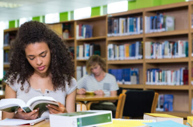 Estudante universitária sentada em uma biblioteca, lendo um livro sobre uma mesa com cadernos, livros e materiais de estudo. Ao fundo, estantes repletas de livros e outro estudante estudando reforçam o ambiente acadêmico. A cena representa a rotina de estudos na faculdade, com foco, concentração e organização.