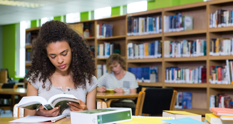 Estudante universitária sentada em uma biblioteca, lendo um livro sobre uma mesa com cadernos, livros e materiais de estudo. Ao fundo, estantes repletas de livros e outro estudante estudando reforçam o ambiente acadêmico. A cena representa a rotina de estudos na faculdade, com foco, concentração e organização.