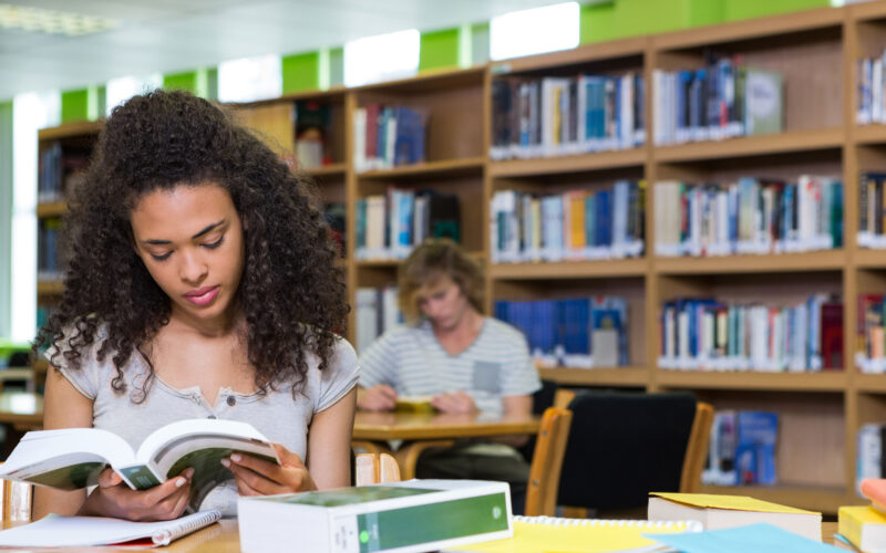 Estudante universitária sentada em uma biblioteca, lendo um livro sobre uma mesa com cadernos, livros e materiais de estudo. Ao fundo, estantes repletas de livros e outro estudante estudando reforçam o ambiente acadêmico. A cena representa a rotina de estudos na faculdade, com foco, concentração e organização.