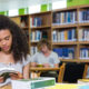 Estudante universitária sentada em uma biblioteca, lendo um livro sobre uma mesa com cadernos, livros e materiais de estudo. Ao fundo, estantes repletas de livros e outro estudante estudando reforçam o ambiente acadêmico. A cena representa a rotina de estudos na faculdade, com foco, concentração e organização.