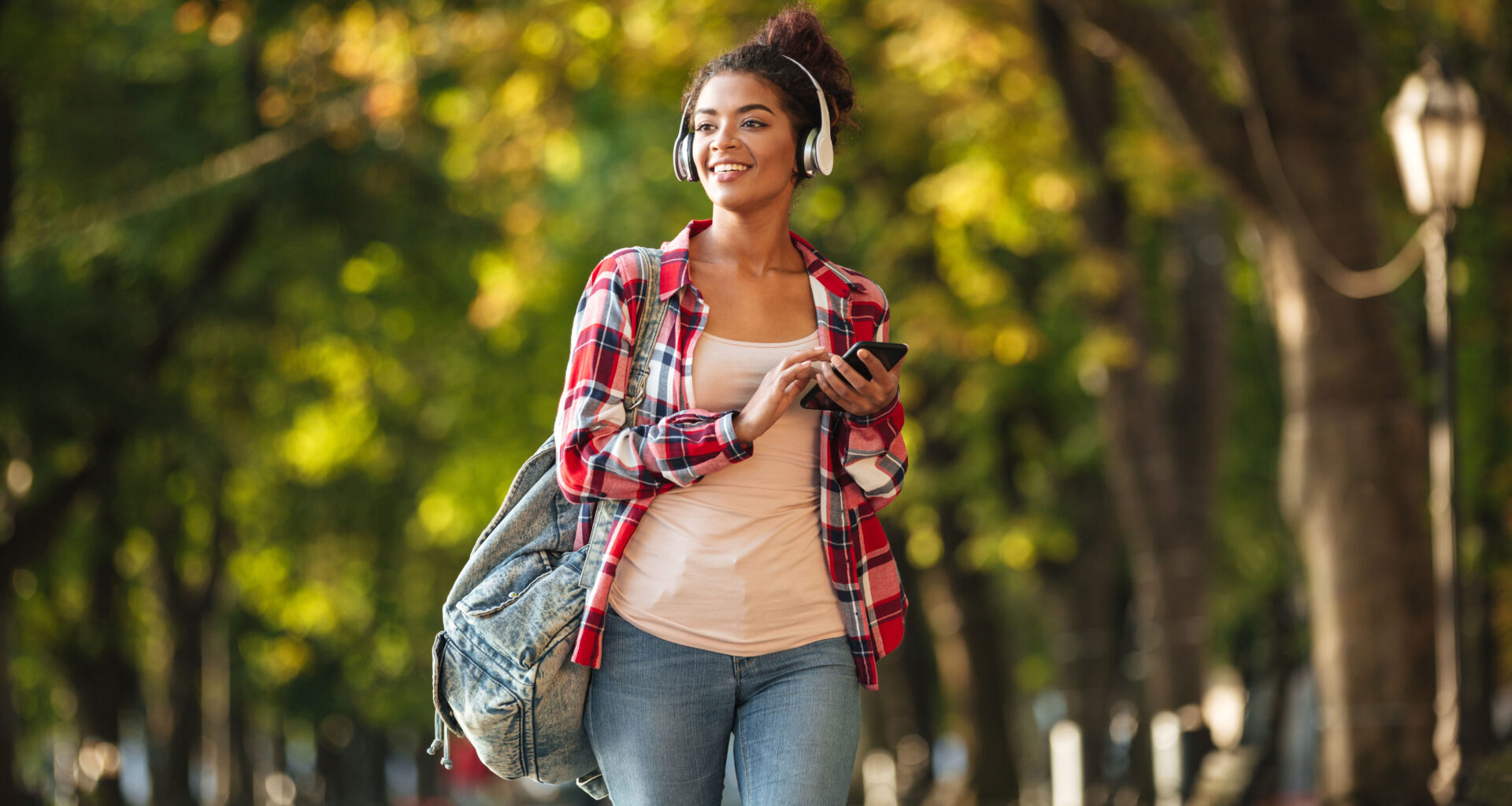 Estudante universitária caminhando por um parque arborizado durante o dia, usando fones de ouvido, mochila nas costas e segurando um celular. A jovem sorri enquanto observa o ambiente ao seu redor, transmitindo uma sensação de descanso, bem-estar e aproveitamento das férias da faculdade para cuidar da saúde, aprender e viver novas experiências.