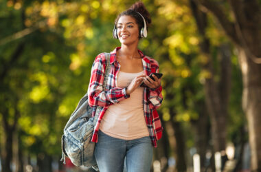 Estudante universitária caminhando por um parque arborizado durante o dia, usando fones de ouvido, mochila nas costas e segurando um celular. A jovem sorri enquanto observa o ambiente ao seu redor, transmitindo uma sensação de descanso, bem-estar e aproveitamento das férias da faculdade para cuidar da saúde, aprender e viver novas experiências.