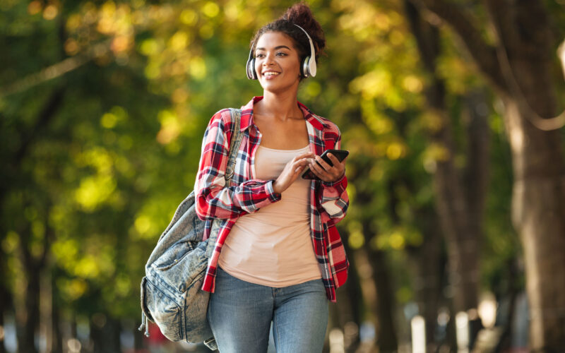 Estudante universitária caminhando por um parque arborizado durante o dia, usando fones de ouvido, mochila nas costas e segurando um celular. A jovem sorri enquanto observa o ambiente ao seu redor, transmitindo uma sensação de descanso, bem-estar e aproveitamento das férias da faculdade para cuidar da saúde, aprender e viver novas experiências.