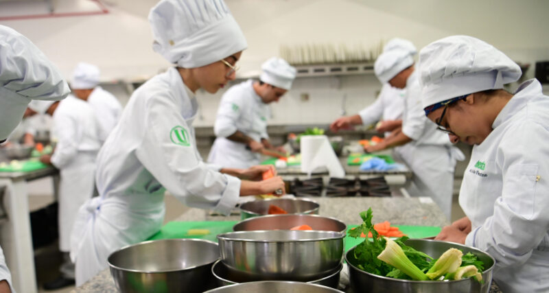 Estudantes da faculdade de Gastronomia da Unoeste participam de uma aula prática em laboratório de cozinha, preparando ingredientes, utilizando utensílios profissionais e desenvolvendo técnicas culinárias essenciais para a formação de futuros chefs de cozinha e personal chefs.