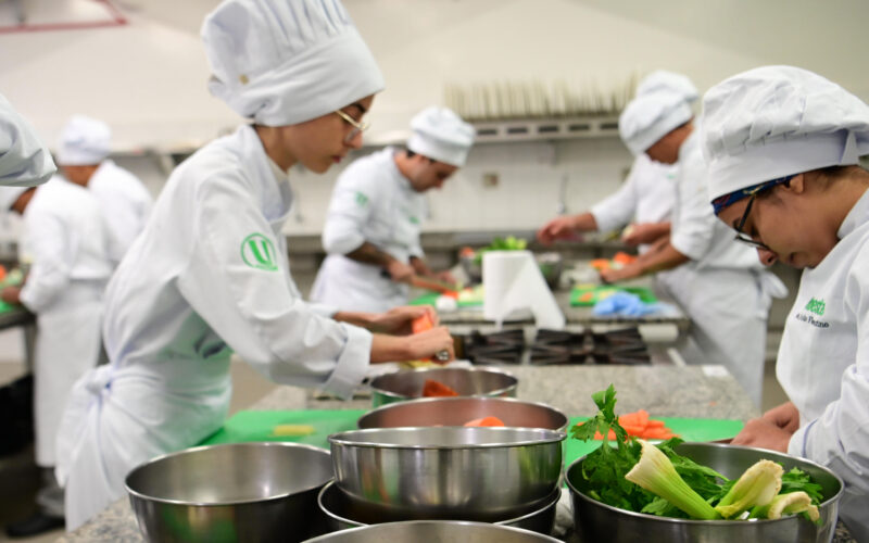 Estudantes da faculdade de Gastronomia da Unoeste participam de uma aula prática em laboratório de cozinha, preparando ingredientes, utilizando utensílios profissionais e desenvolvendo técnicas culinárias essenciais para a formação de futuros chefs de cozinha e personal chefs.