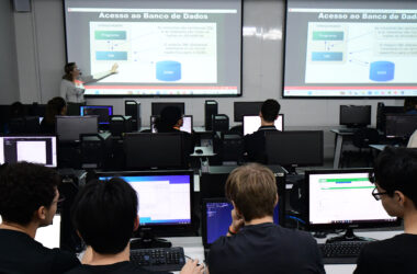 Professora ministra aula sobre acesso a banco de dados em laboratório de informática da Unoeste, enquanto estudantes de Sistemas de Informação acompanham a explicação e desenvolvem atividades práticas nos computadores.