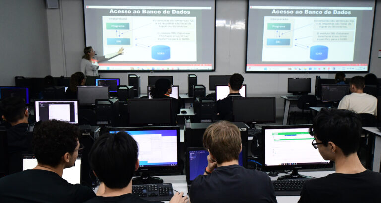 Professora ministra aula sobre acesso a banco de dados em laboratório de informática da Unoeste, enquanto estudantes de Sistemas de Informação acompanham a explicação e desenvolvem atividades práticas nos computadores.