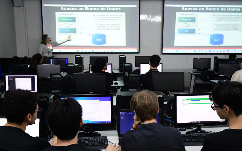 Professora ministra aula sobre acesso a banco de dados em laboratório de informática da Unoeste, enquanto estudantes de Sistemas de Informação acompanham a explicação e desenvolvem atividades práticas nos computadores.
