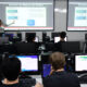 Professora ministra aula sobre acesso a banco de dados em laboratório de informática da Unoeste, enquanto estudantes de Sistemas de Informação acompanham a explicação e desenvolvem atividades práticas nos computadores.
