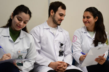 studantes de Medicina da Unoeste, usando jalecos, realizam atividade acadêmica em grupo durante aula prática, registrando informações em pranchetas e discutindo casos clínicos em ambiente de aprendizagem colaborativa.