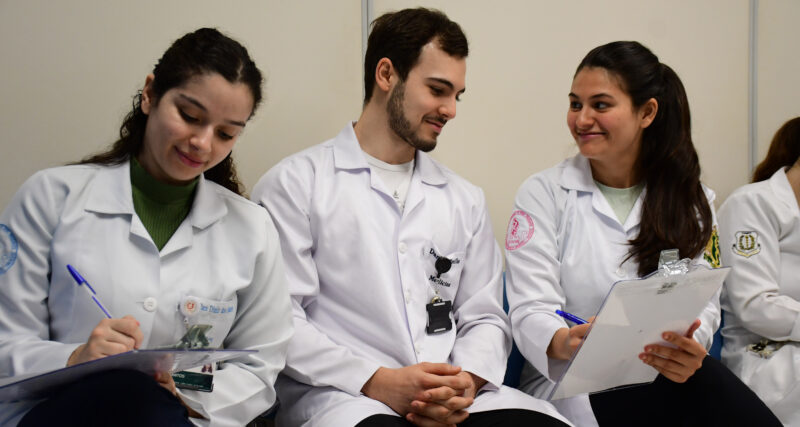 studantes de Medicina da Unoeste, usando jalecos, realizam atividade acadêmica em grupo durante aula prática, registrando informações em pranchetas e discutindo casos clínicos em ambiente de aprendizagem colaborativa.