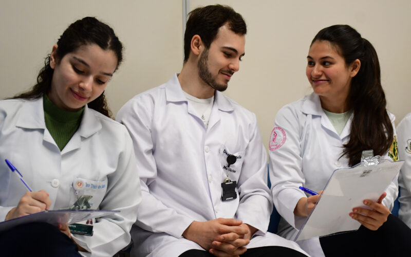 studantes de Medicina da Unoeste, usando jalecos, realizam atividade acadêmica em grupo durante aula prática, registrando informações em pranchetas e discutindo casos clínicos em ambiente de aprendizagem colaborativa.