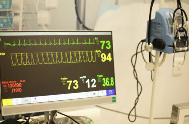 Monitor multiparamétrico exibe frequência cardíaca, pressão arterial, saturação de oxigênio e outros sinais vitais em ambiente de prática médica.