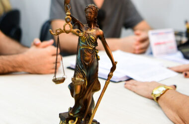 Estátua da Justiça com balança em primeiro plano, enquanto estudantes participam de atividade acadêmica com documentos sobre uma mesa ao fundo.