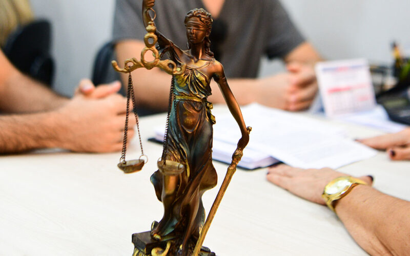 Estátua da Justiça com balança em primeiro plano, enquanto estudantes participam de atividade acadêmica com documentos sobre uma mesa ao fundo.