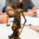 Estátua da Justiça com balança em primeiro plano, enquanto estudantes participam de atividade acadêmica com documentos sobre uma mesa ao fundo.