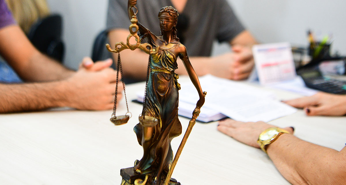 Estátua da Justiça com balança em primeiro plano, enquanto estudantes participam de atividade acadêmica com documentos sobre uma mesa ao fundo.