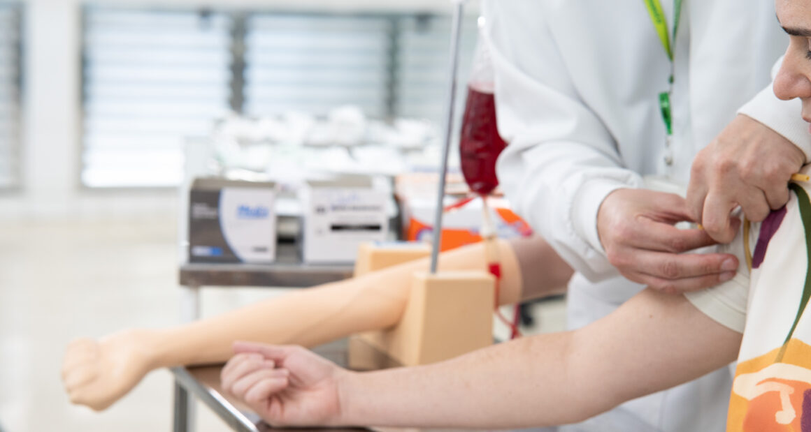 Profissional de saúde prepara o braço de uma pessoa para doação de sangue em ambiente de coleta, com bolsa de sangue e equipamentos utilizados no procedimento ao fundo.