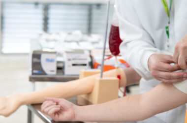 Profissional de saúde prepara o braço de uma pessoa para doação de sangue em ambiente de coleta, com bolsa de sangue e equipamentos utilizados no procedimento ao fundo.