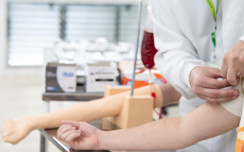 Profissional de saúde prepara o braço de uma pessoa para doação de sangue em ambiente de coleta, com bolsa de sangue e equipamentos utilizados no procedimento ao fundo.
