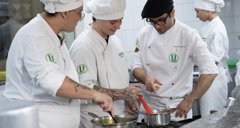Estudantes de Gastronomia da Unoeste preparam receitas durante aula prática em cozinha profissional, acompanhando técnicas de preparo dos alimentos.