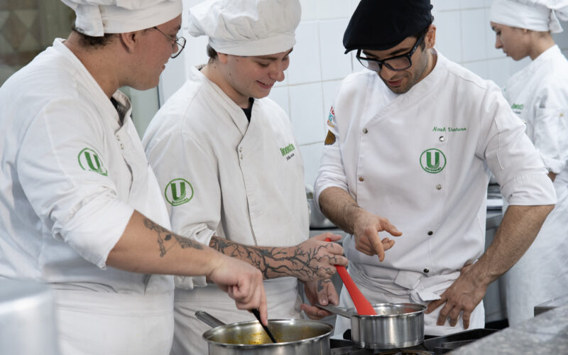 Estudantes de Gastronomia da Unoeste preparam receitas durante aula prática em cozinha profissional, acompanhando técnicas de preparo dos alimentos.