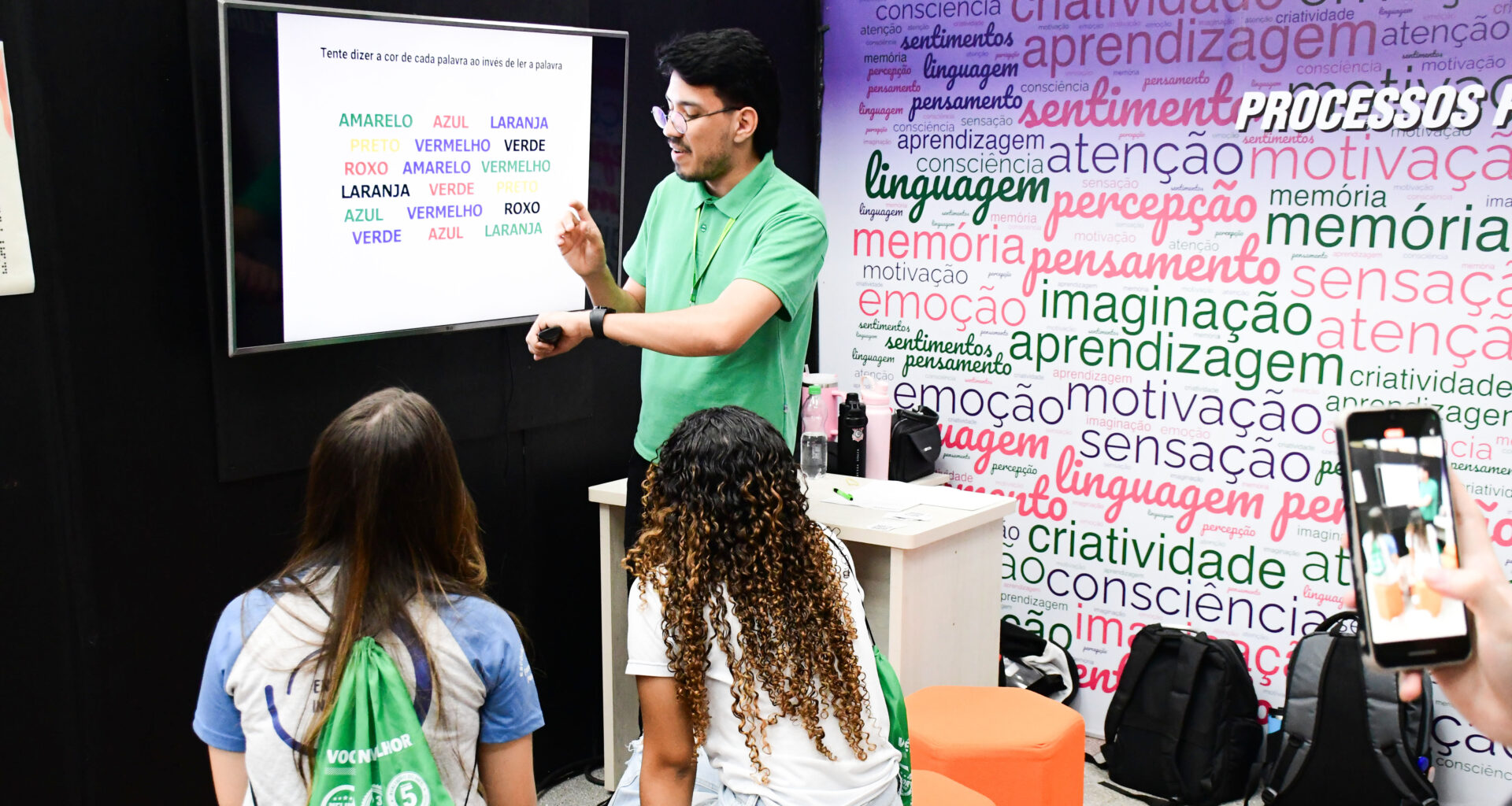Professor apresenta atividade sobre atenção e percepção em uma tela digital enquanto estudantes acompanham a explicação em sala de aula, em ambiente com palavras relacionadas à aprendizagem, memória, criatividade, linguagem e emoção.