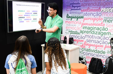 Professor apresenta atividade sobre atenção e percepção em uma tela digital enquanto estudantes acompanham a explicação em sala de aula, em ambiente com palavras relacionadas à aprendizagem, memória, criatividade, linguagem e emoção.