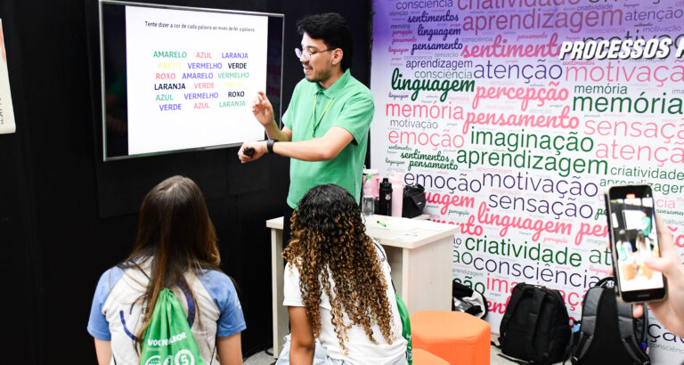 Professor apresenta atividade sobre atenção e percepção em uma tela digital enquanto estudantes acompanham a explicação em sala de aula, em ambiente com palavras relacionadas à aprendizagem, memória, criatividade, linguagem e emoção.