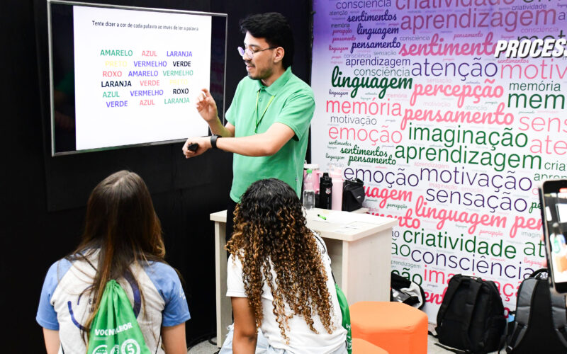 Professor apresenta atividade sobre atenção e percepção em uma tela digital enquanto estudantes acompanham a explicação em sala de aula, em ambiente com palavras relacionadas à aprendizagem, memória, criatividade, linguagem e emoção.