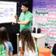 Professor apresenta atividade sobre atenção e percepção em uma tela digital enquanto estudantes acompanham a explicação em sala de aula, em ambiente com palavras relacionadas à aprendizagem, memória, criatividade, linguagem e emoção.