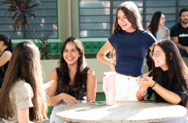 Grupo de estudantes universitárias conversando e sorrindo ao redor de uma mesa em área de convivência do campus, durante um momento de interação e integração na rotina da faculdade.