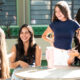 Grupo de estudantes universitárias conversando e sorrindo ao redor de uma mesa em área de convivência do campus, durante um momento de interação e integração na rotina da faculdade.