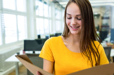 Jovem profissional sorrindo enquanto analisa documentos em uma pasta, em um escritório com computadores e mesas de trabalho ao fundo, representando possibilidades de atuação para bacharel em Direito sem OAB.