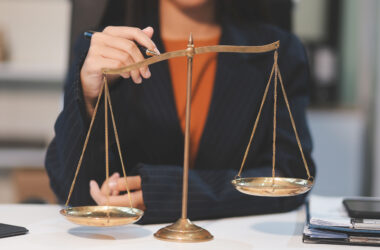 Balança da Justiça em destaque sobre uma mesa de escritório, enquanto uma pessoa em traje social interage com o instrumento, representando as diferentes carreiras na área do Direito.