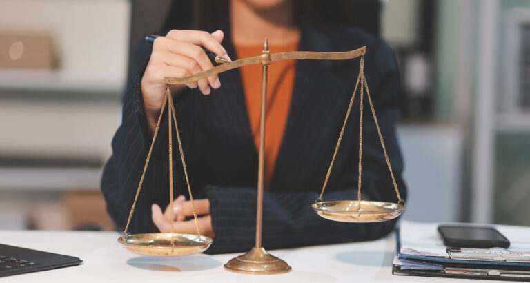 Balança da Justiça em destaque sobre uma mesa de escritório, enquanto uma pessoa em traje social interage com o instrumento, representando as diferentes carreiras na área do Direito.