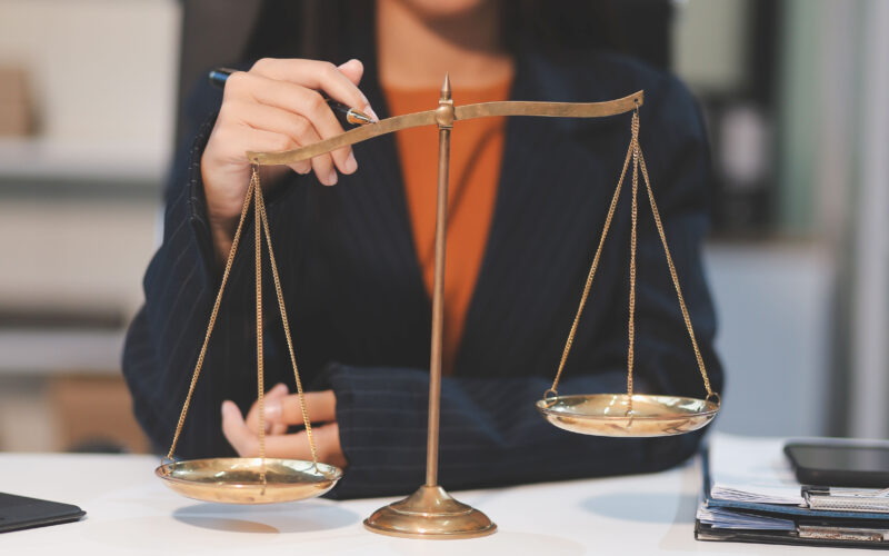 Balança da Justiça em destaque sobre uma mesa de escritório, enquanto uma pessoa em traje social interage com o instrumento, representando as diferentes carreiras na área do Direito.