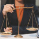 Balança da Justiça em destaque sobre uma mesa de escritório, enquanto uma pessoa em traje social interage com o instrumento, representando as diferentes carreiras na área do Direito.