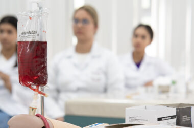 Bolsa de sangue em primeiro plano durante aula prática com estudantes da área da Saúde ao fundo.
