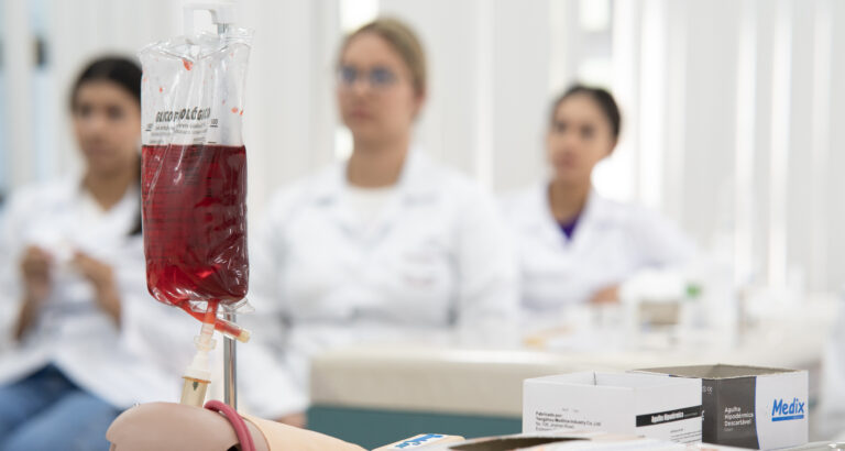 Bolsa de sangue em primeiro plano durante aula prática com estudantes da área da Saúde ao fundo.