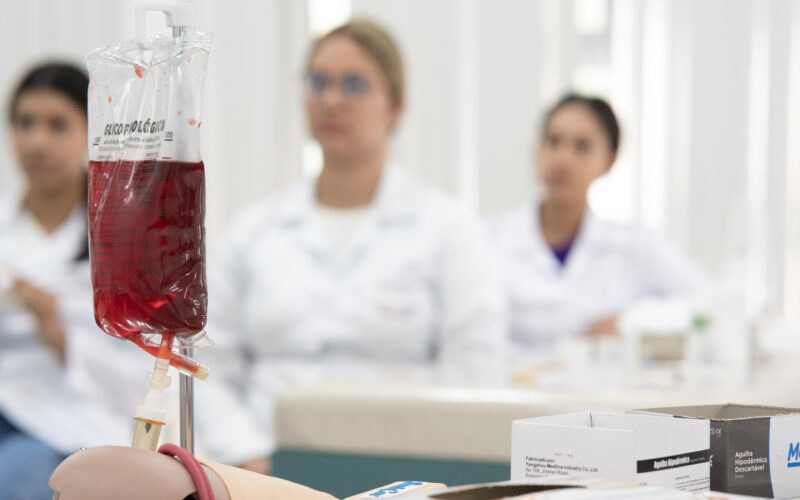 Bolsa de sangue em primeiro plano durante aula prática com estudantes da área da Saúde ao fundo.