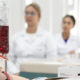 Bolsa de sangue em primeiro plano durante aula prática com estudantes da área da Saúde ao fundo.