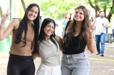 Três estudantes universitárias sorrindo juntas em área externa arborizada do campus, duas delas fazendo sinal de paz com as mãos, enquanto outras pessoas circulam ao fundo.