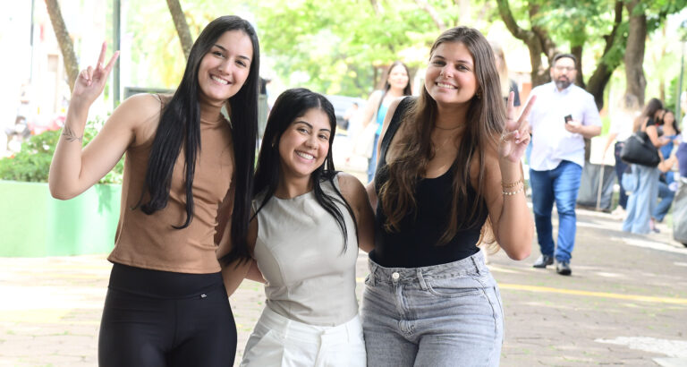 Três estudantes universitárias sorrindo juntas em área externa arborizada do campus, duas delas fazendo sinal de paz com as mãos, enquanto outras pessoas circulam ao fundo.