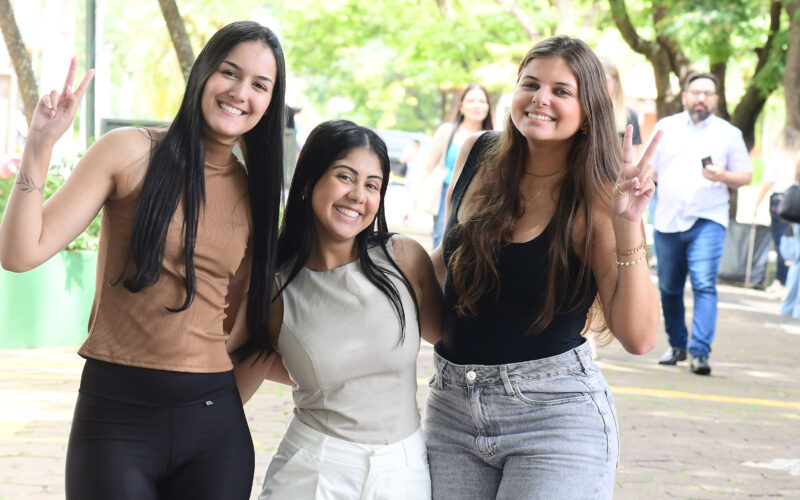 Três estudantes universitárias sorrindo juntas em área externa arborizada do campus, duas delas fazendo sinal de paz com as mãos, enquanto outras pessoas circulam ao fundo.
