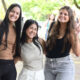 Três estudantes universitárias sorrindo juntas em área externa arborizada do campus, duas delas fazendo sinal de paz com as mãos, enquanto outras pessoas circulam ao fundo.
