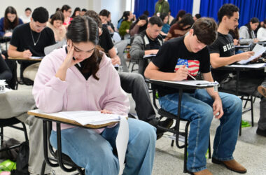 Estudantes realizam prova em sala de aula, sentados individualmente em carteiras enquanto respondem às questões impressas.