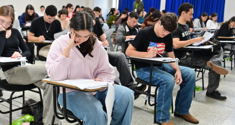 Estudantes realizam prova em sala de aula, sentados individualmente em carteiras enquanto respondem às questões impressas.