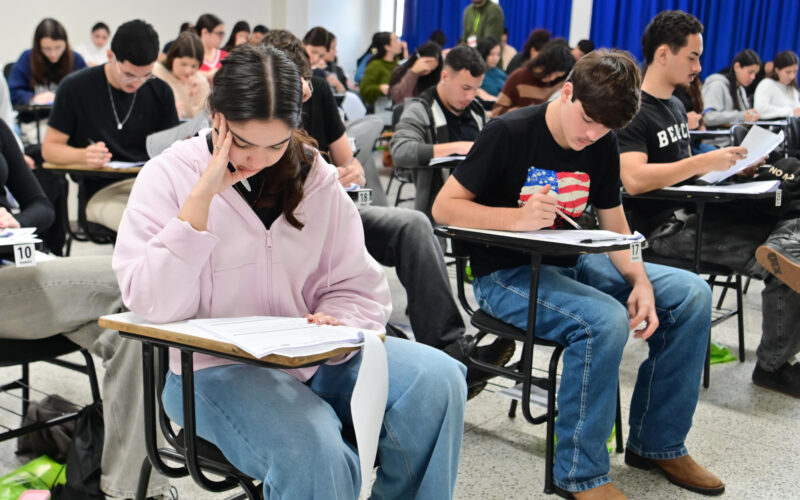 Estudantes realizam prova em sala de aula, sentados individualmente em carteiras enquanto respondem às questões impressas.